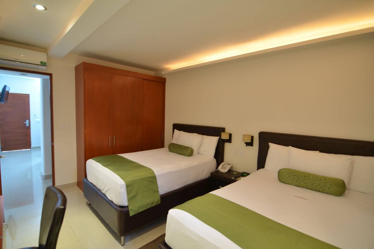 Habitación de hotel con dos camas, cada una con almohada verde y ropa de cama blanca. Se observa un armario de madera y una mesita de noche con un teléfono.