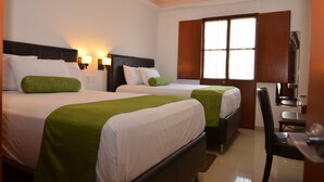 Een hotelkamer met twee bedden, elk met een groene sprei en witte lakens, een bureau met stoel en een groot raam met houten luiken.