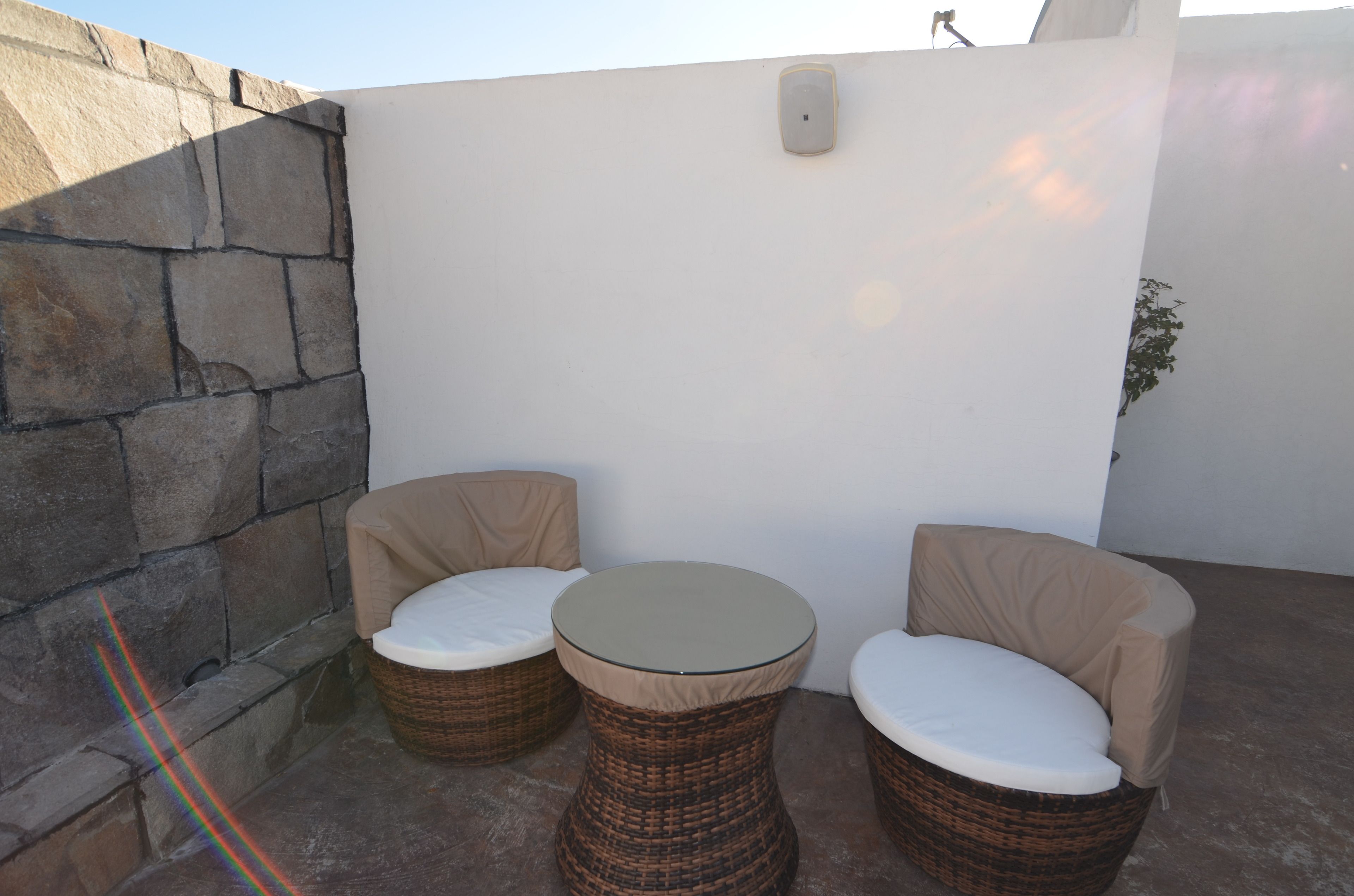 A patio area with two wicker chairs and a small round table, a stone wall, and a white wall with a light fixture.
