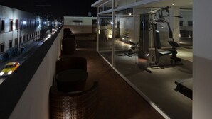 Een balkon met fitnessapparatuur, een rieten stoel en een glazen wand met uitzicht op een straat in de nacht.