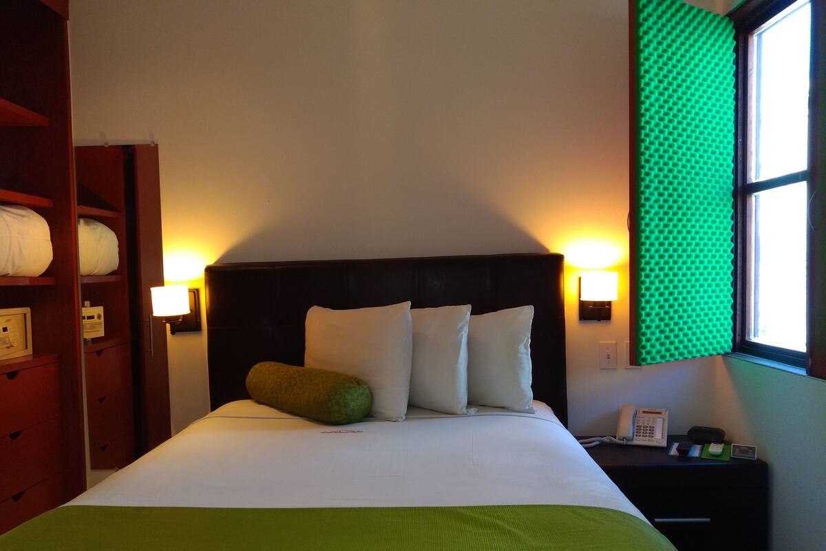 Una cama bien hecha con almohadas y una manta verde, una lámpara de noche, un teléfono y una ventana con persianas verdes.