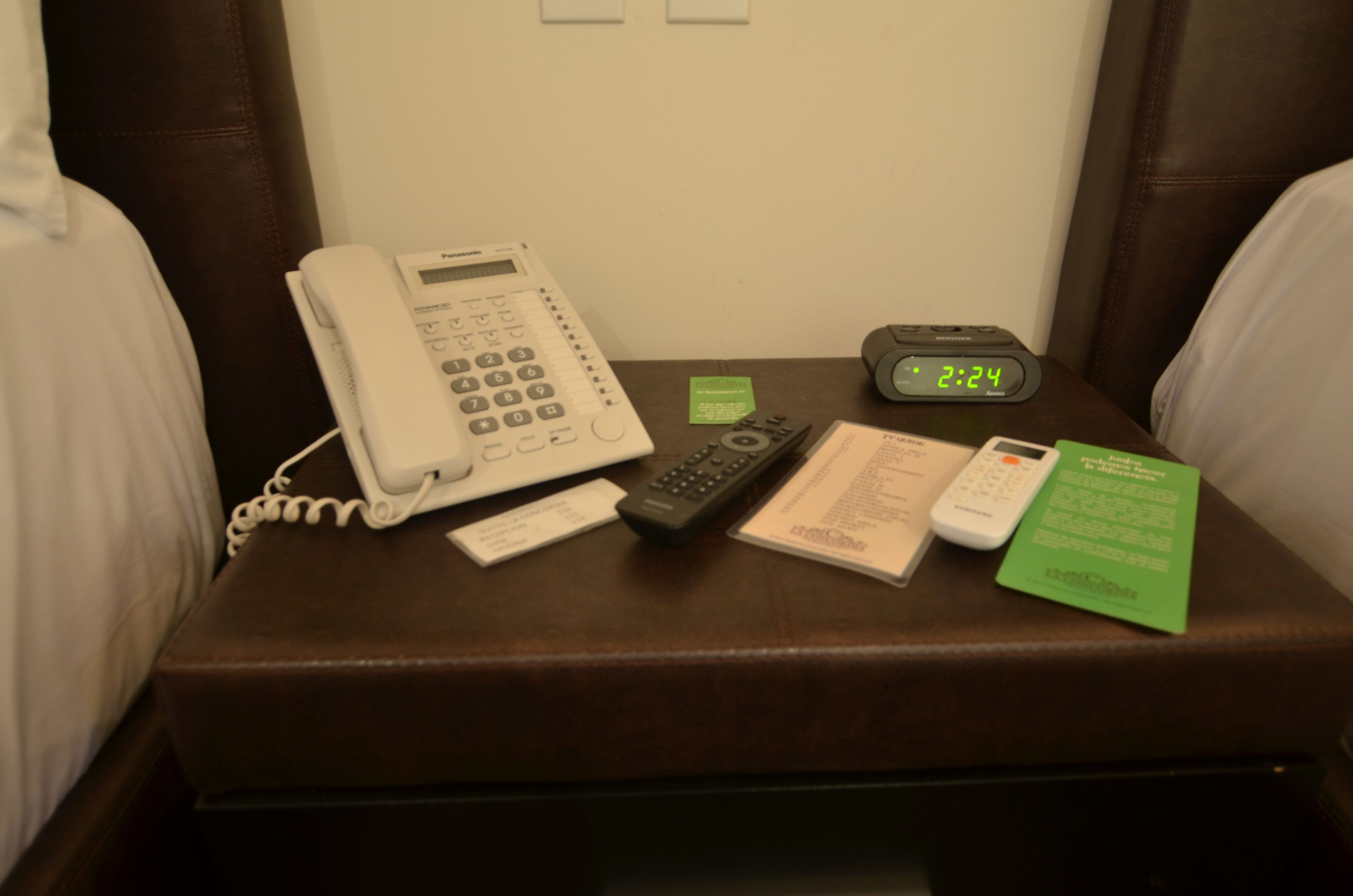 A hotel room with a brown leather armchair, a white telephone, a digital clock, a remote control, and a green card on the armchair.