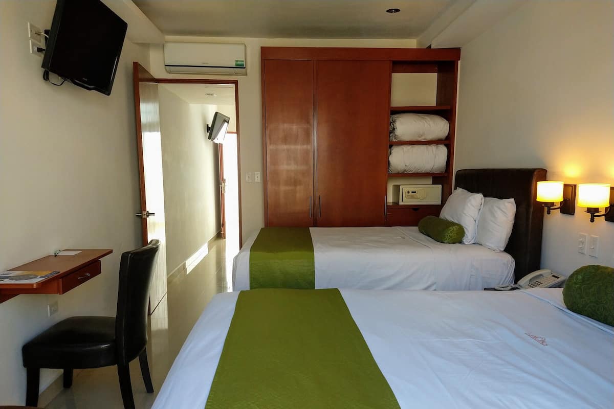 Una habitación de hotel con dos camas, un escritorio, una silla, un televisor, aire acondicionado, un armario y una lámpara de noche.