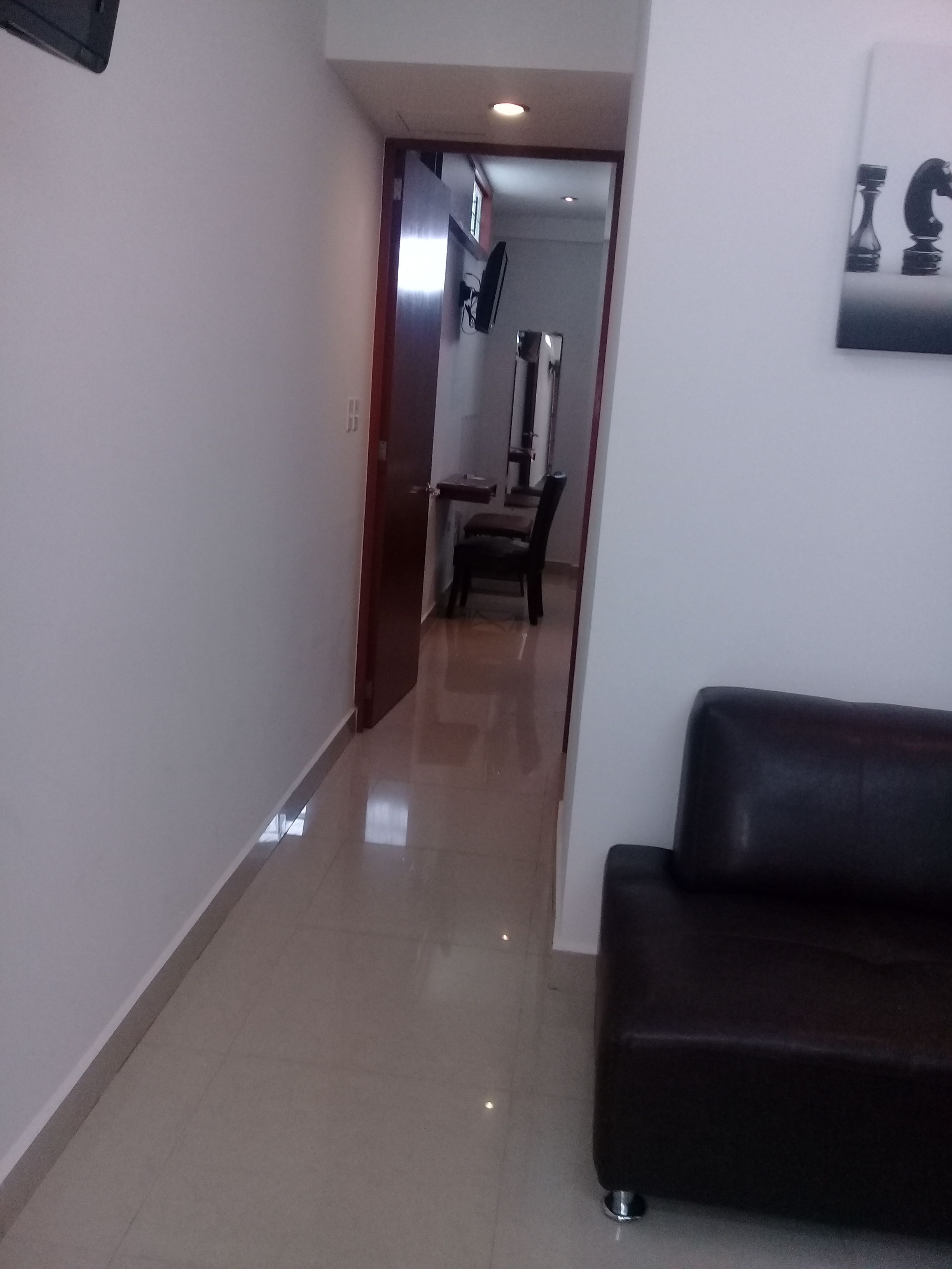 Un pasillo con un sofá de cuero negro, suelo de baldosas y una puerta que da a una habitación con un espejo y una silla.