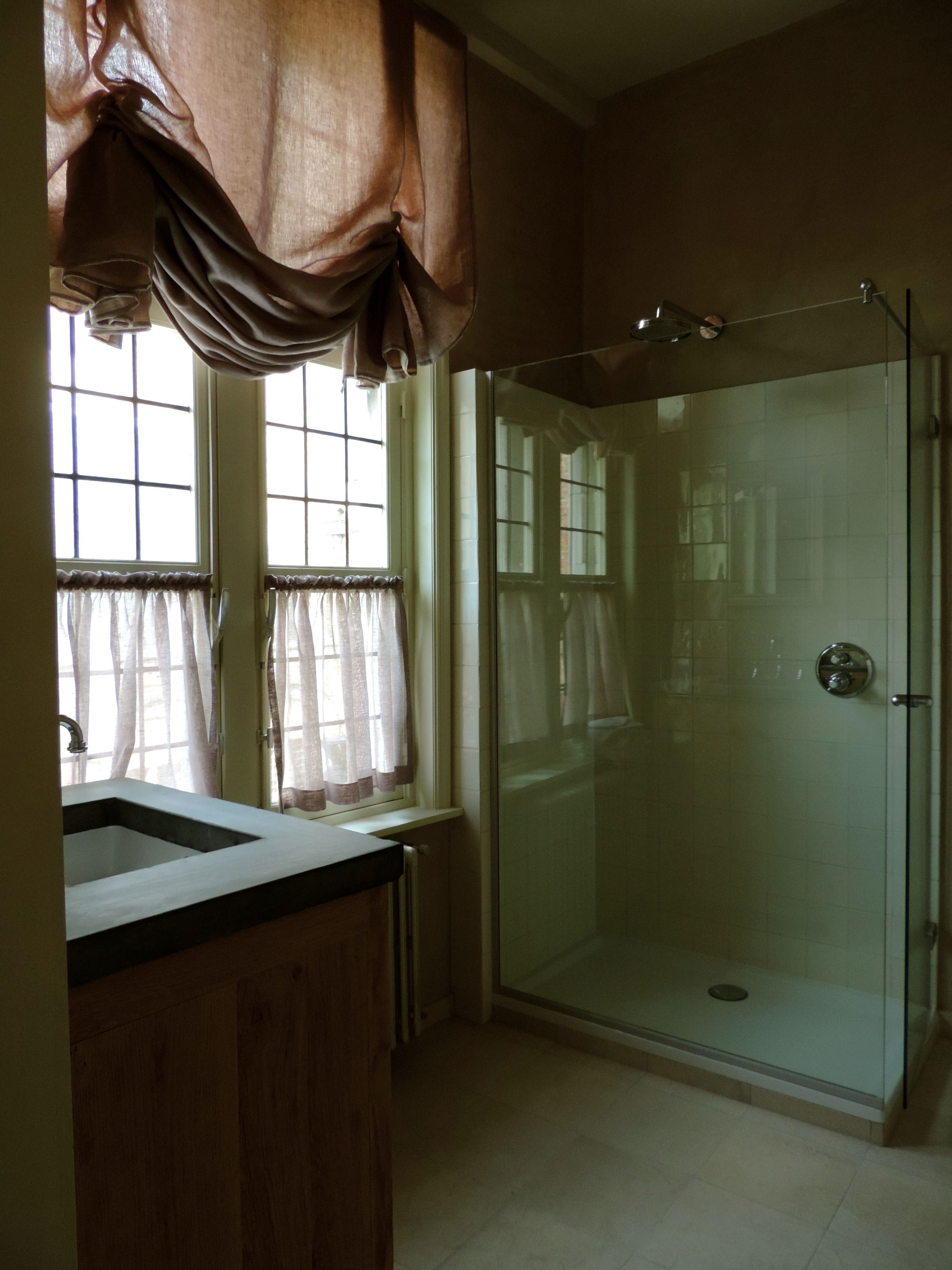 A bathroom with a glass shower enclosure, a wooden vanity, and a window with curtains.