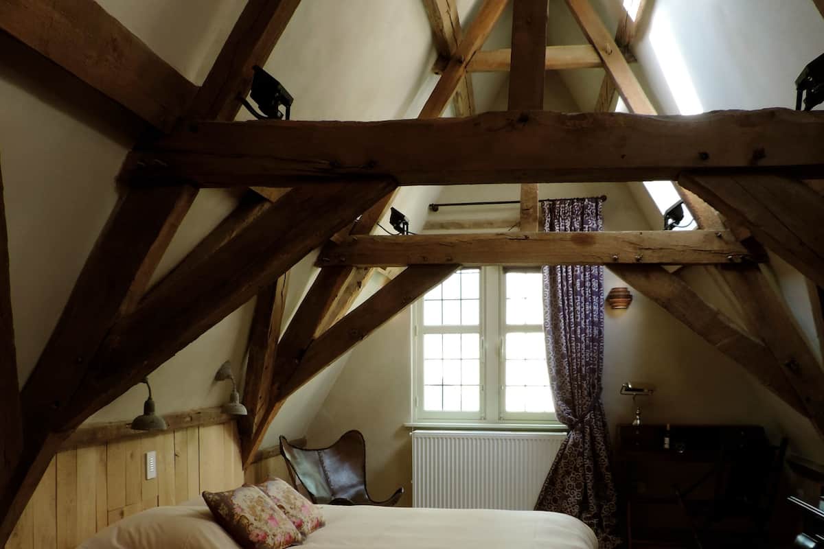 Un dormitorio con techo inclinado, vigas de madera, una cama con almohadas, una silla, un escritorio y una ventana con cortinas.