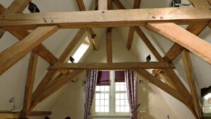 Un dormitorio con techo inclinado, vigas de madera, una cama con colcha morada, un escritorio con silla y una ventana con cortinas.