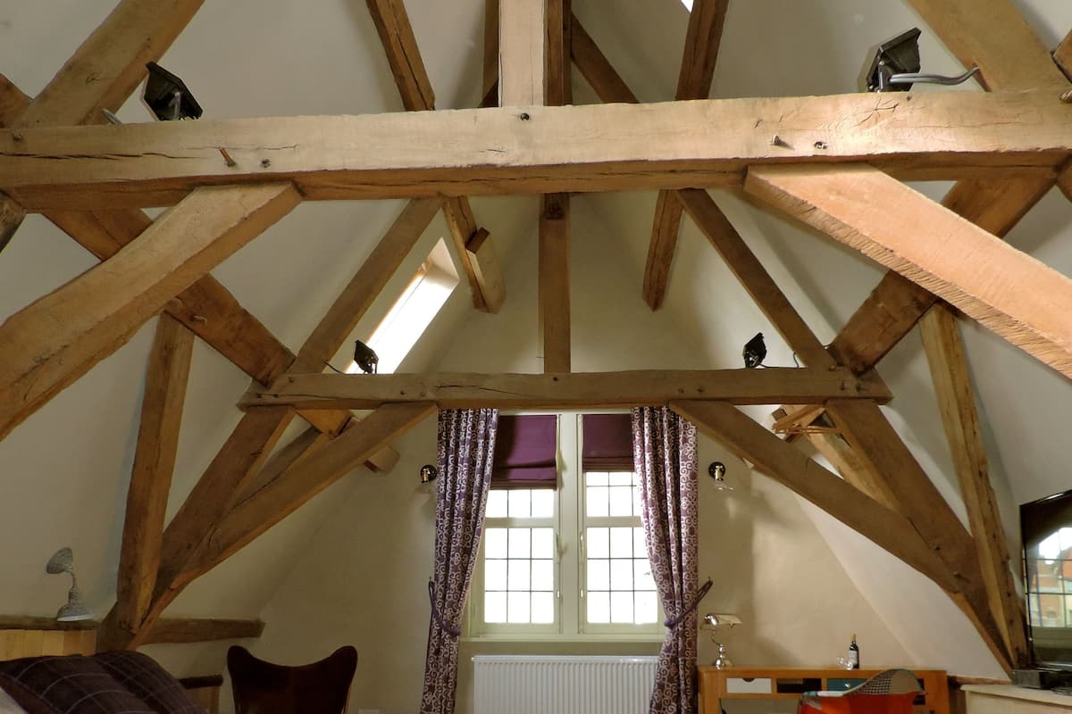 Un dormitorio con techo inclinado, vigas de madera, una cama con colcha morada, un escritorio con silla y una ventana con cortinas.