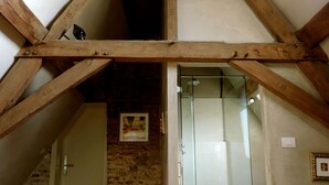 Una habitación con techo inclinado, vigas de madera, mampara de cristal para la ducha y una puerta que da a otra habitación.