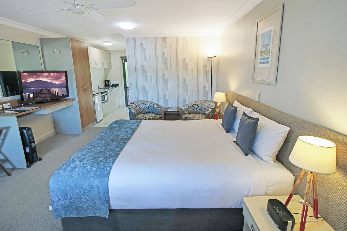 A hotel room with a bed, bedside table, lamp, TV, desk, and kitchenette.