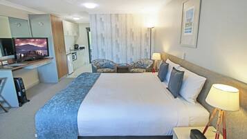 Quarto de hotel com cama, mesa de cabeceira, luminária, TV, escrivaninha e cozinha compacta.