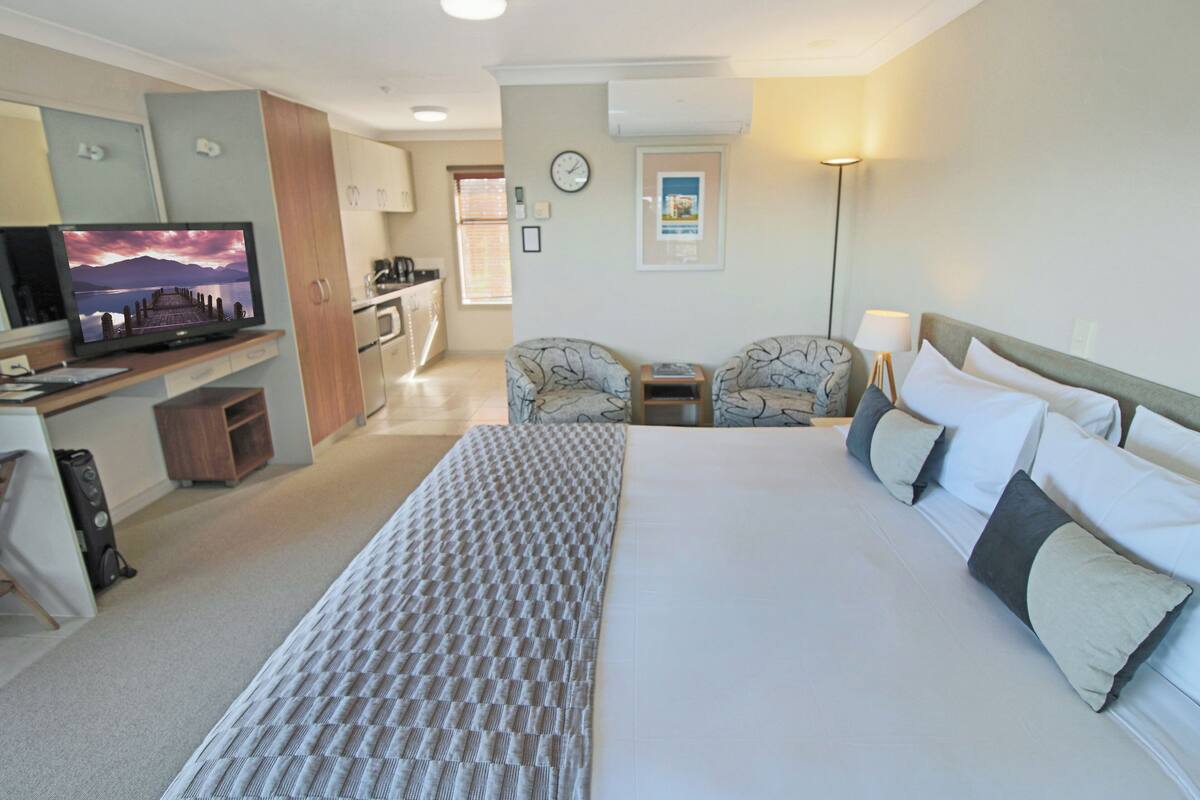 A hotel room with a large bed, a desk with a TV, a kitchenette, and a seating area.