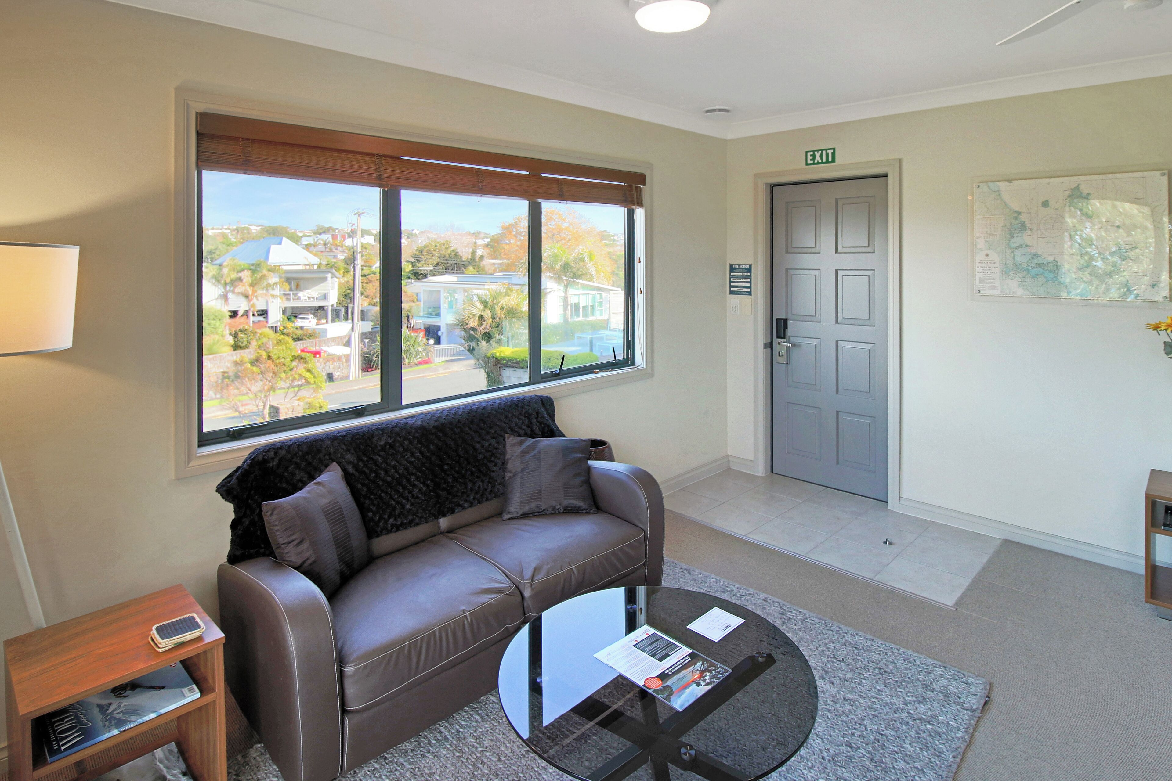Une chambre d’hôtel moderne avec un canapé en cuir, une table basse en verre, une petite table d’appoint, une fenêtre avec des stores et une porte marquée « SORTIE ».