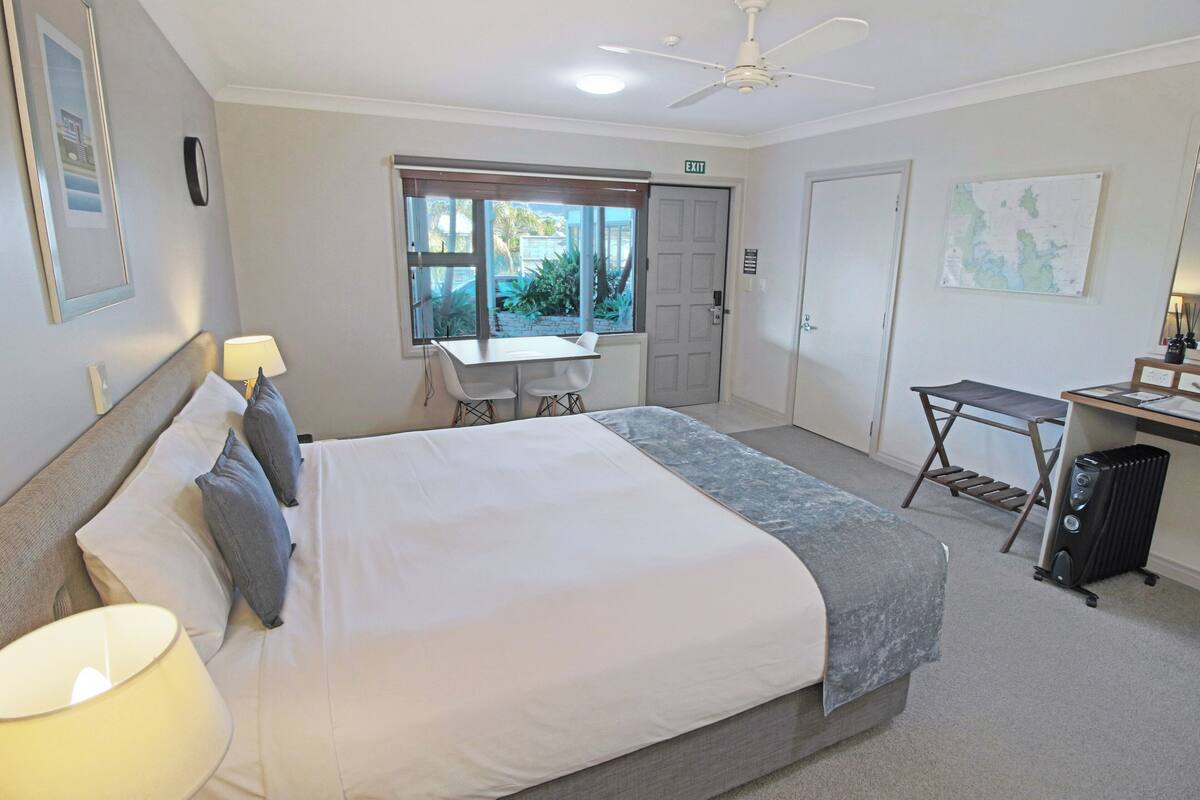 A hotel room with a bed, bedside table, lamp, desk, chair, and a view of greenery outside.