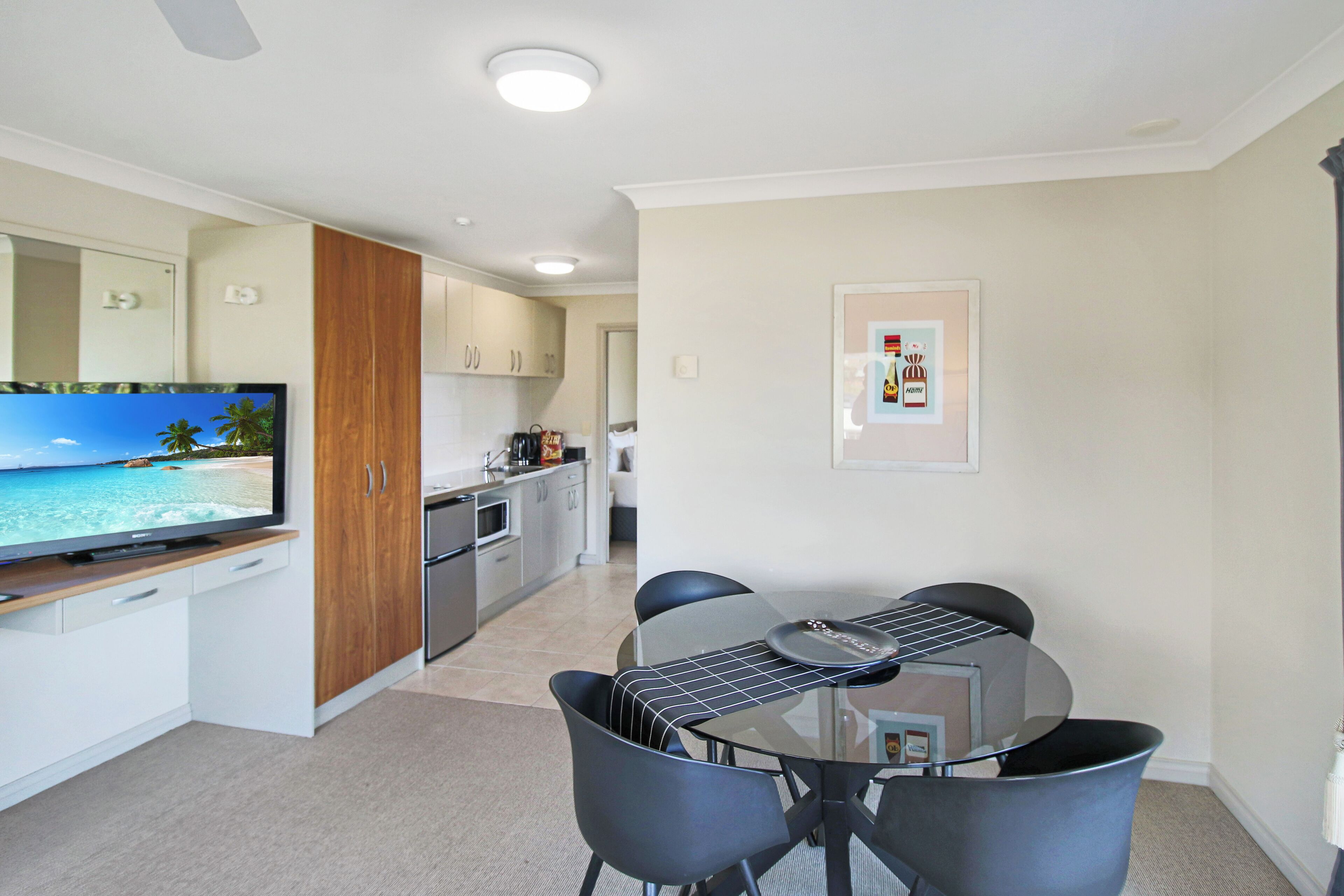 A modern kitchen with white cabinets, a wooden cabinet, and a flat-screen TV displaying a beach scene. There is a dining area with a glass table and four chairs.