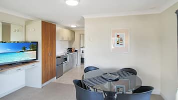 A modern kitchen with white cabinets, a wooden cabinet, and a flat-screen TV displaying a beach scene. There is a dining area with a glass table and four chairs.