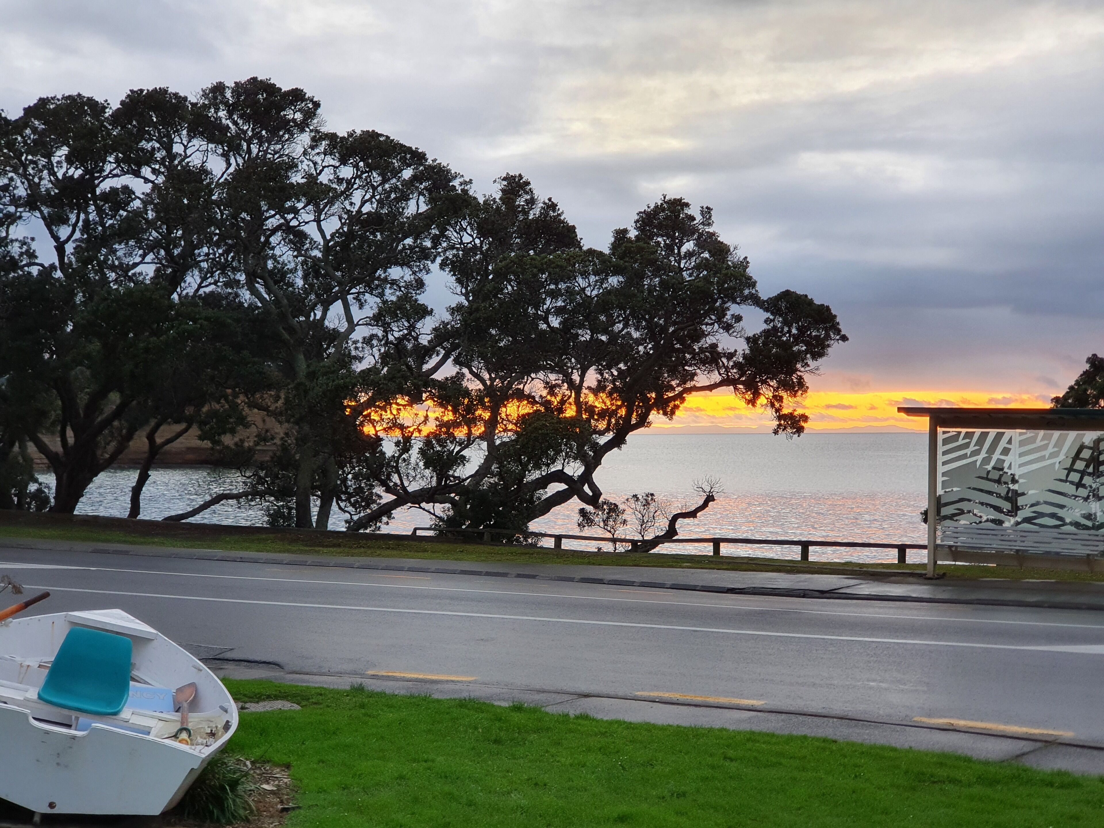 Una carretera costera con un barco estacionado al lado, árboles y un atardecer sobre el agua.