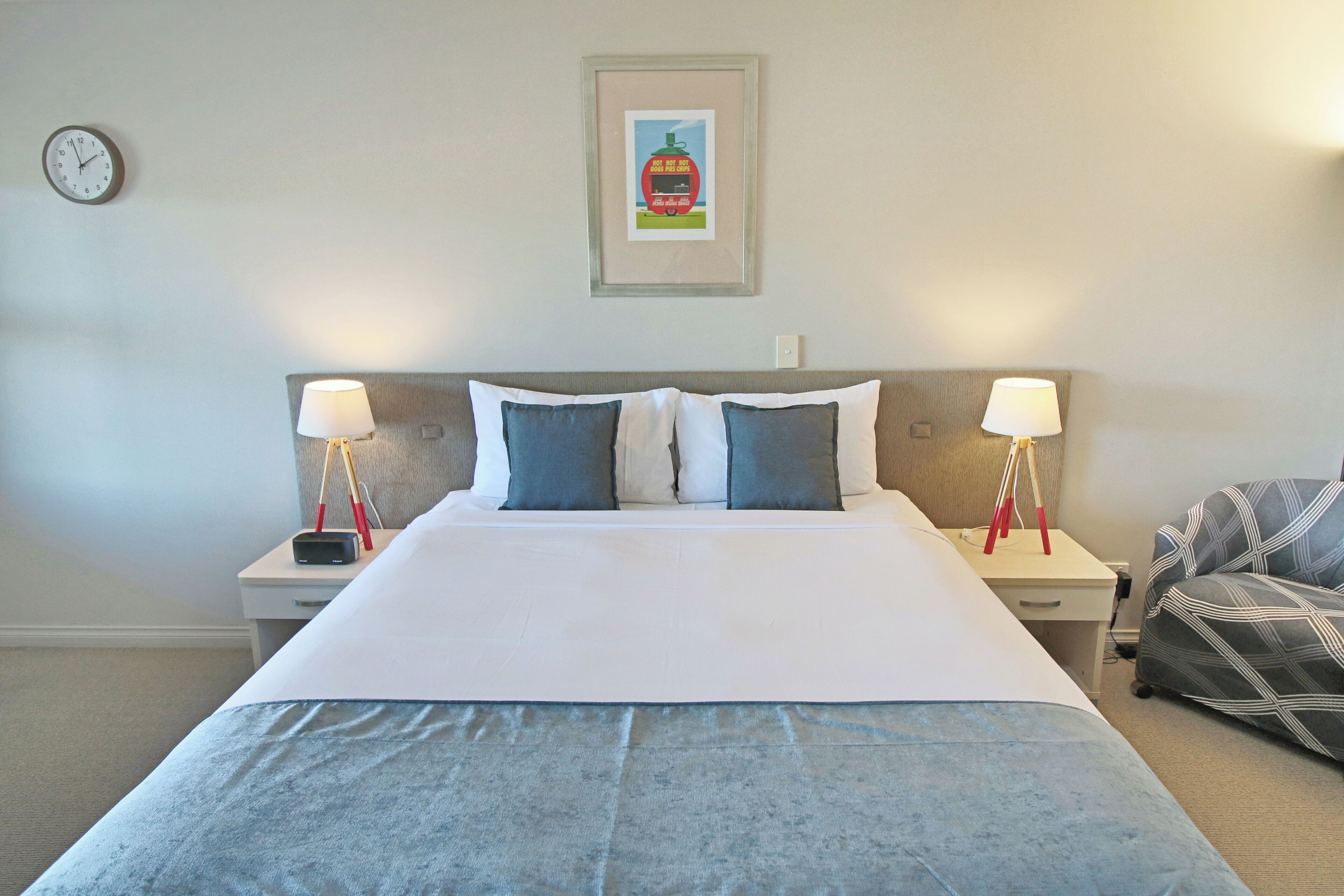 A neatly made bed with a white and blue bedspread, two bedside tables with lamps, a framed picture on the wall, and a clock.