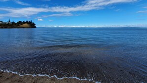 A coastal view with a sandy beach, calm sea, and a distant mountain range under a partly cloudy sky. - Whangaparaoa Lodge Motel (Whangaparaoa)
