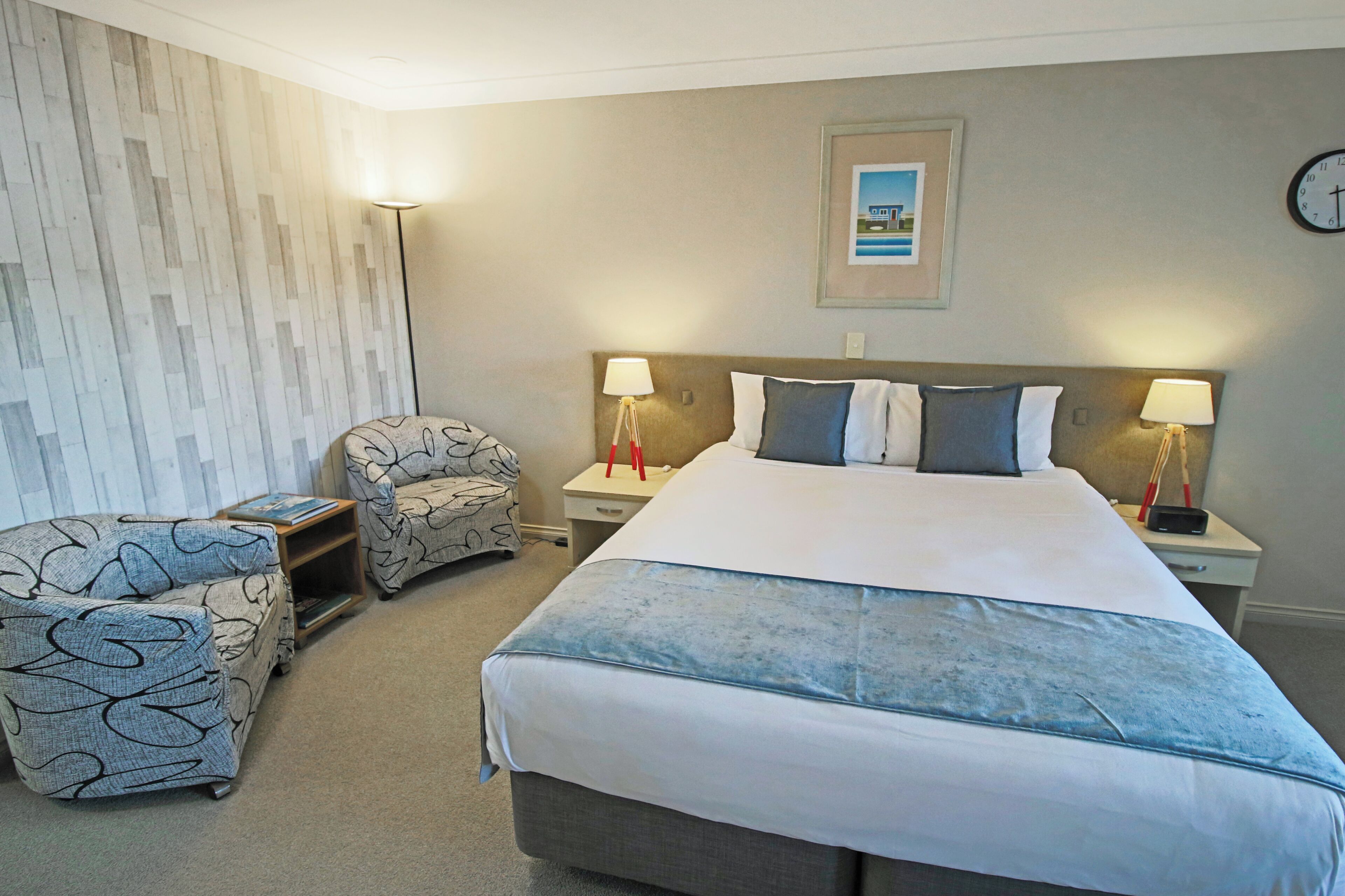 A hotel room with a large bed, two bedside tables with lamps, a chair, and a wall clock.