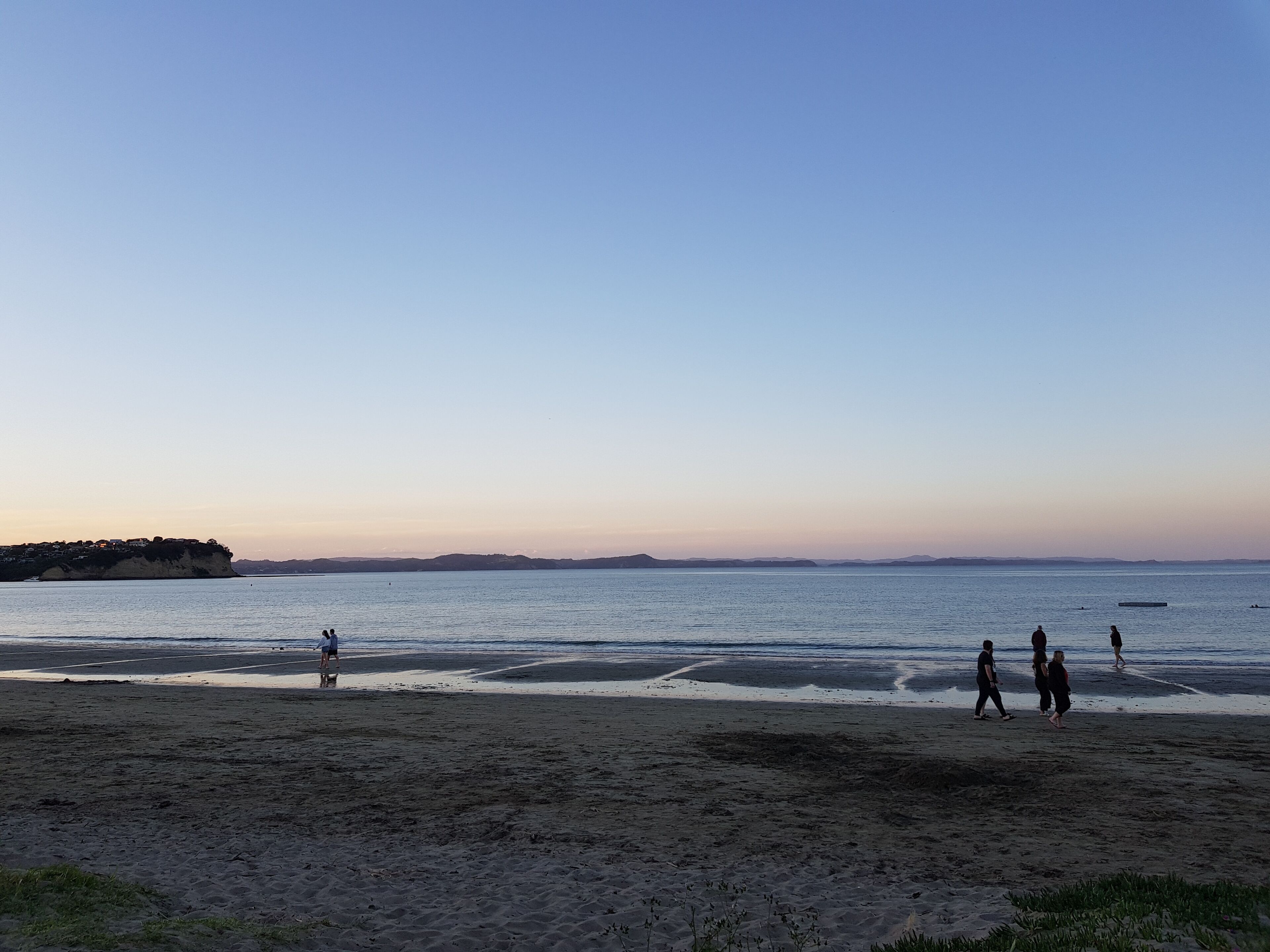 A beach with people walking along the shore, a distant island, and calm waters.