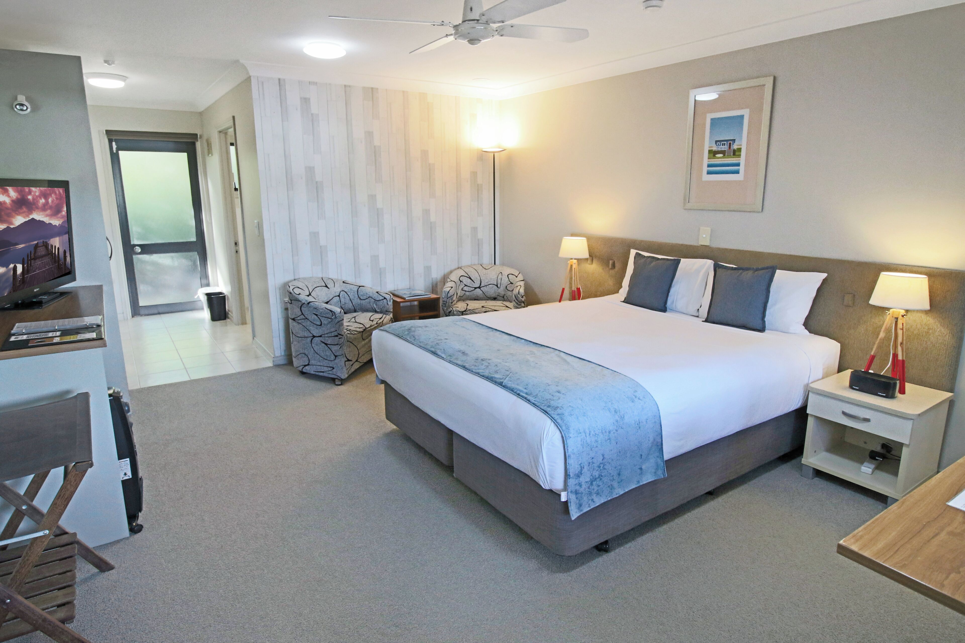 A hotel room with a large bed, two bedside tables, a desk with a chair, a television, and a ceiling fan.