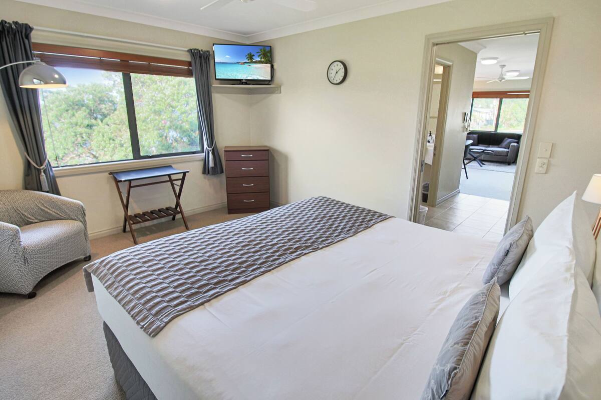 A bedroom with a bed, a desk, a chair, a television, and a clock.