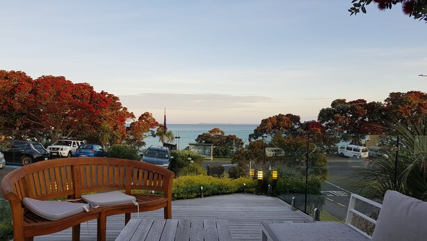 A wooden deck with a bench and a small table, overlooking a parking area and a body of water. - Whangaparaoa Lodge Motel (Whangaparaoa)