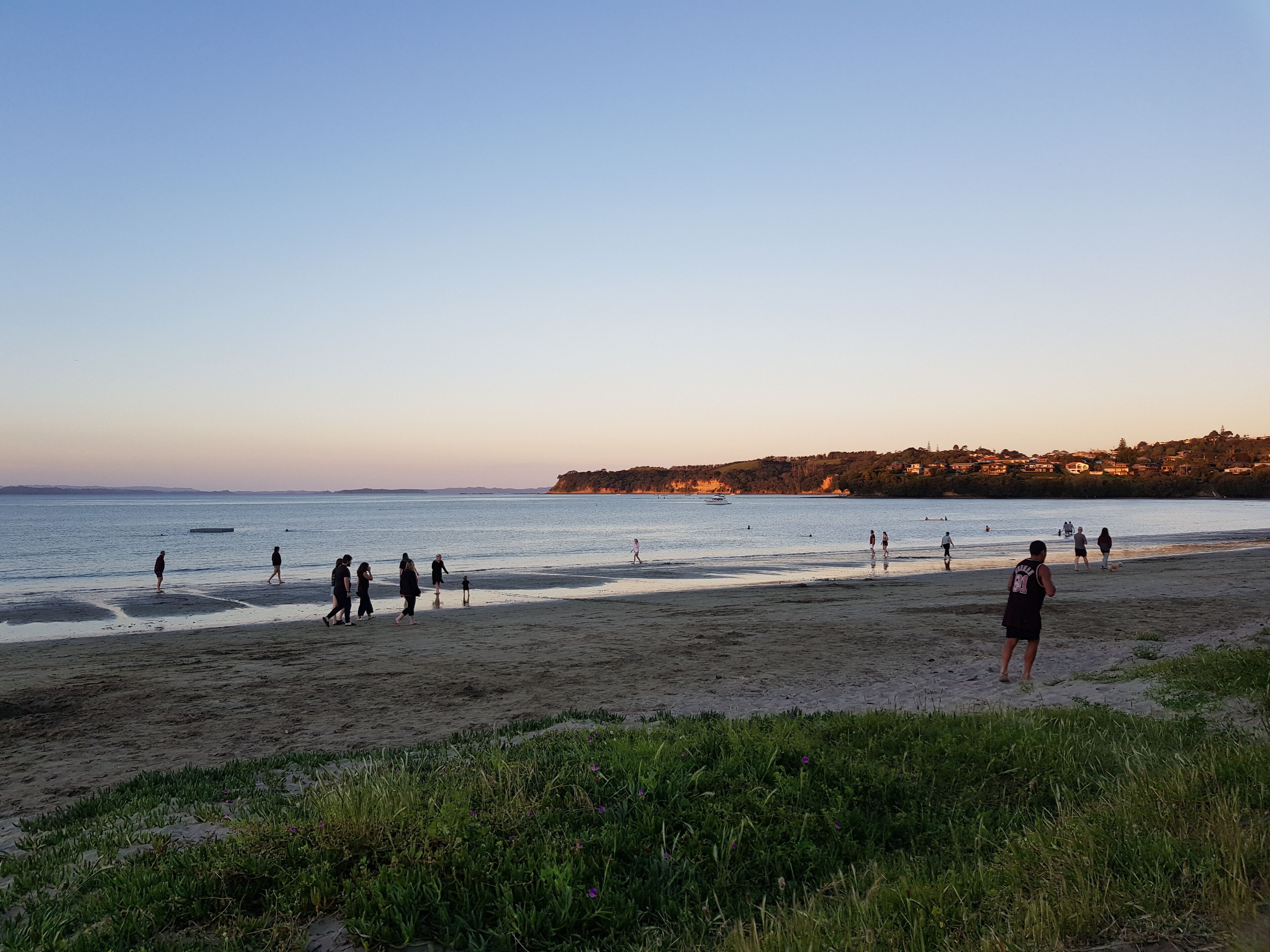 A beach with people walking and playing, a clear sky, and a distant shoreline with buildings.