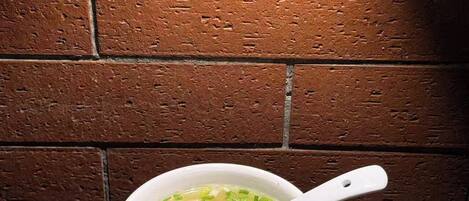 A bowl of soup with a white spoon on a black tray, placed on a wooden table against a brick wall.