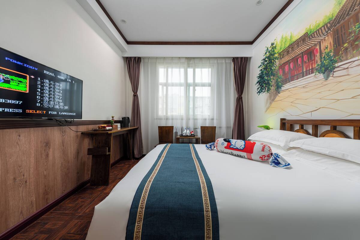 A hotel room with a large bed, a television on the wall, a small table with a chair, and a window with curtains.