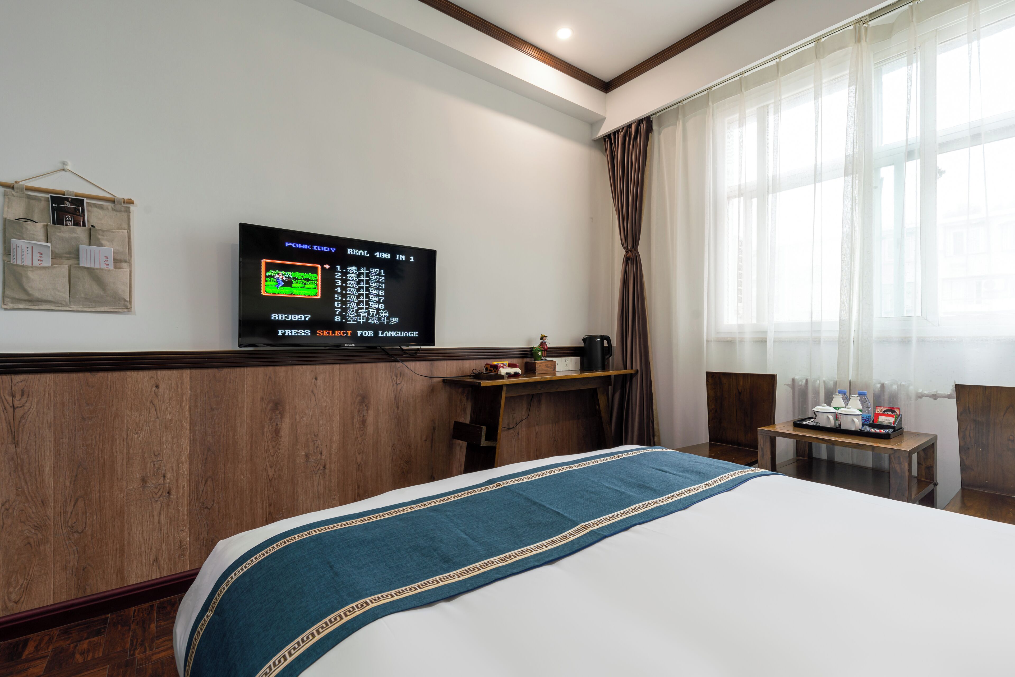A hotel room with a bed, a television mounted on the wall, a wooden headboard, a small table with toiletries, and a window with curtains.