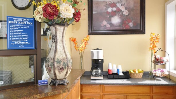 Un'area reception con orologio, un quadro floreale incorniciato, un vaso con fiori, una macchina da caffè e un cartello che indica gli orari di check-in e le regole.