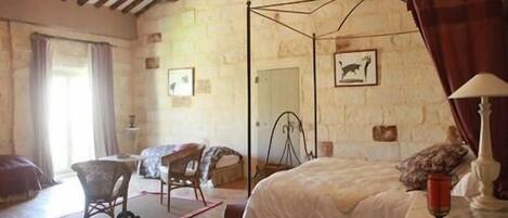 A bedroom with a four-poster bed, a wooden ceiling, stone walls, a small table with a lamp, and a chair.