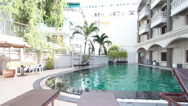 A swimming pool with lounge chairs, a table, and chairs, surrounded by a building with balconies and palm trees. - Honey Lodge (Pattaya)