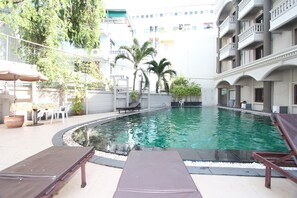 A swimming pool with lounge chairs, a table, and chairs, surrounded by a building with balconies and palm trees.