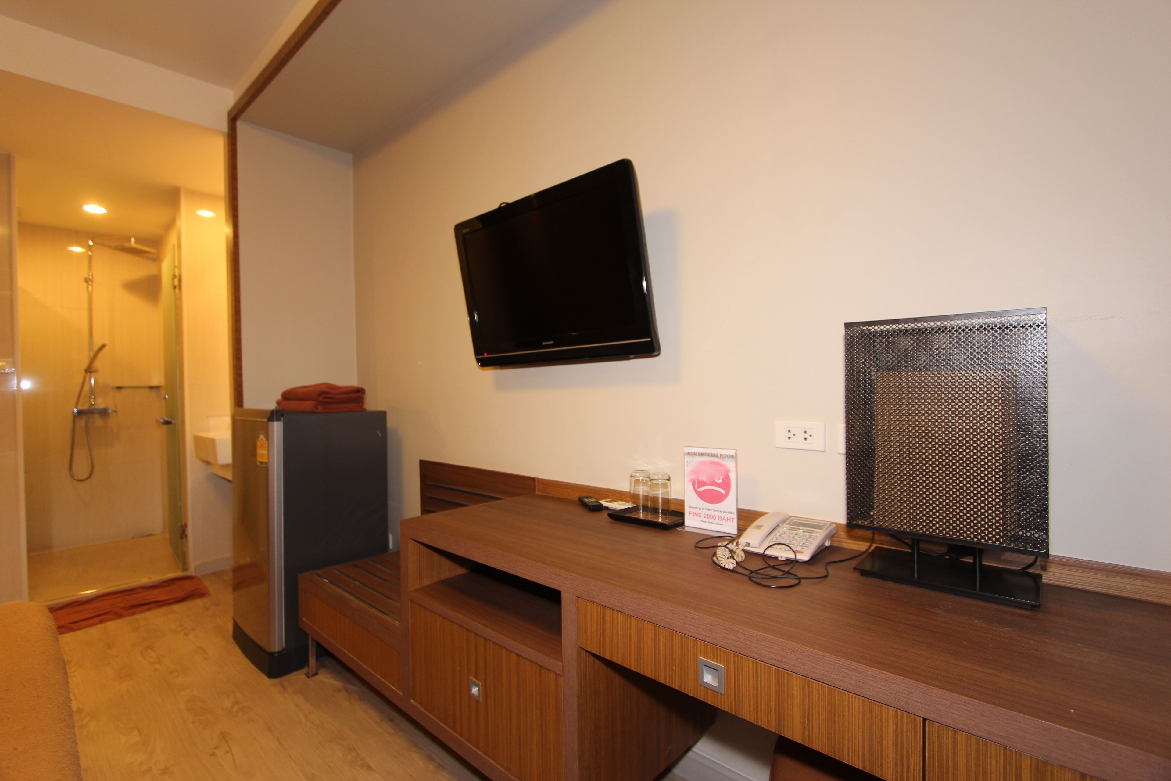 A hotel room with a wooden desk, a flat-screen TV mounted on the wall, a refrigerator, a shower area, and a telephone.