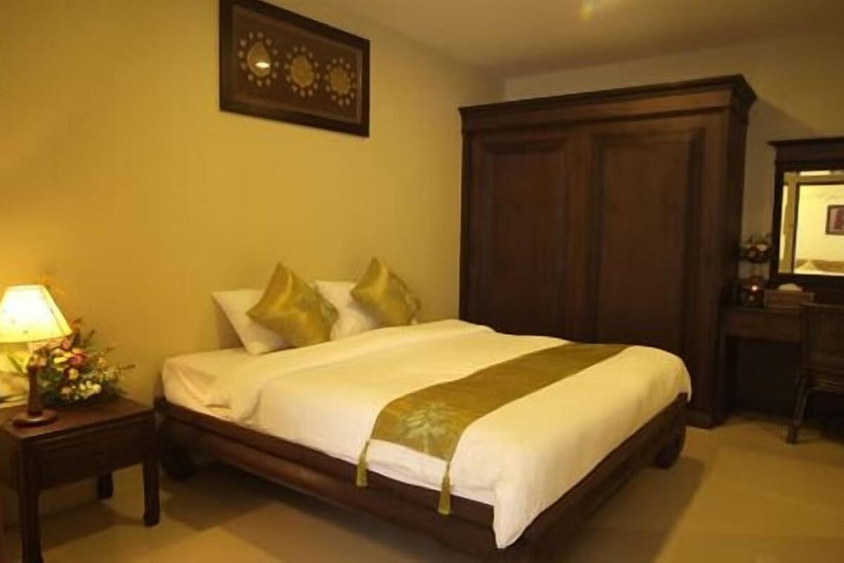 A neatly made bed with a white and gold bedspread, a wooden nightstand with a lamp and flowers, and a large wooden wardrobe.