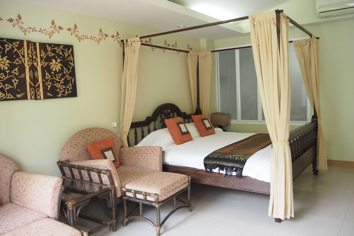 A four-poster bed with beige curtains, a wooden headboard, and a patterned bedspread. There is a chair with a floral pattern and a cushion, and a wall with a decorative panel.