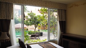 A hotel room with a large window offering a view of a courtyard with a tree, a bench, and a building with a traditional roof. - Villa Thongbura (Pattaya)