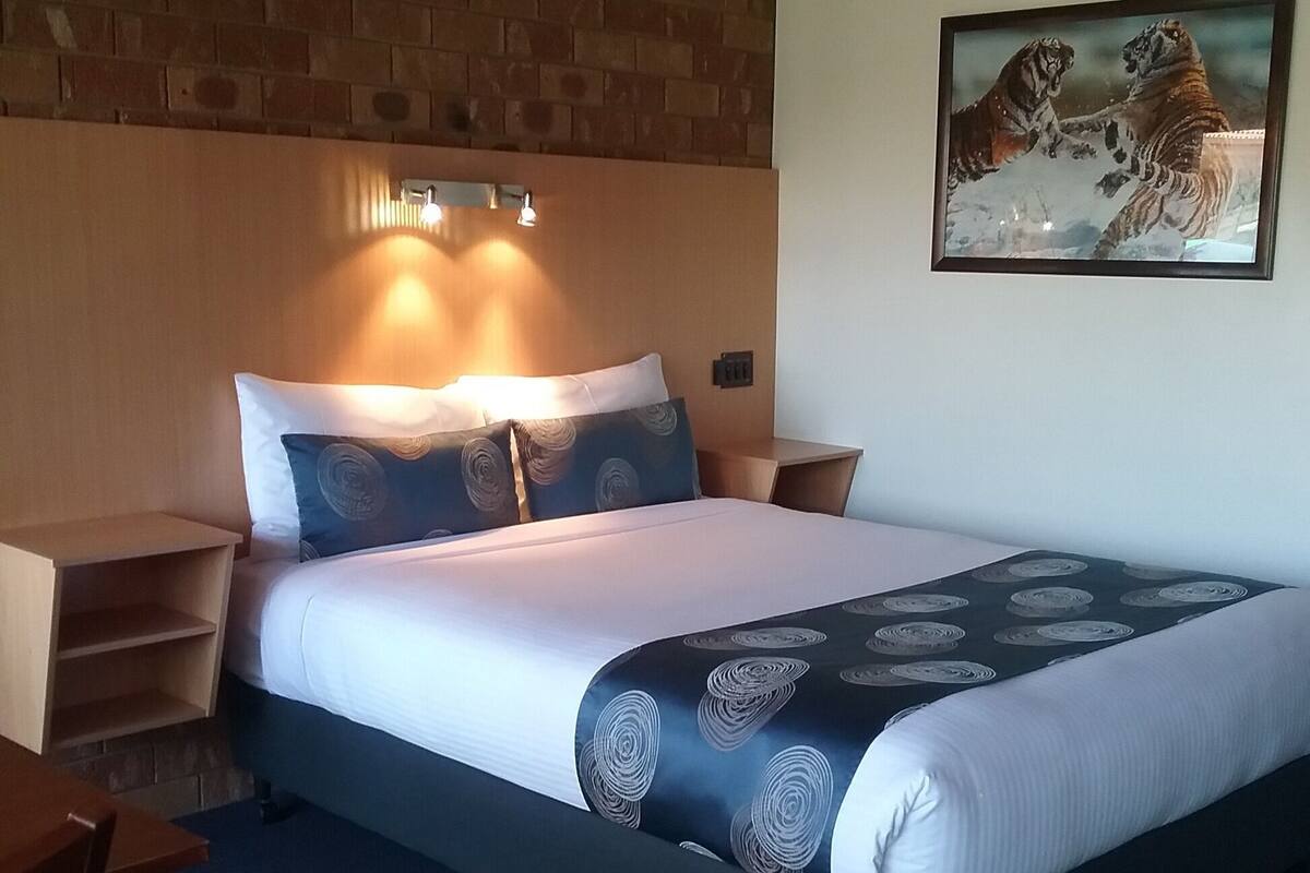 A hotel room with a bed, a wooden headboard, a nightstand, a desk, and a painting on the wall.