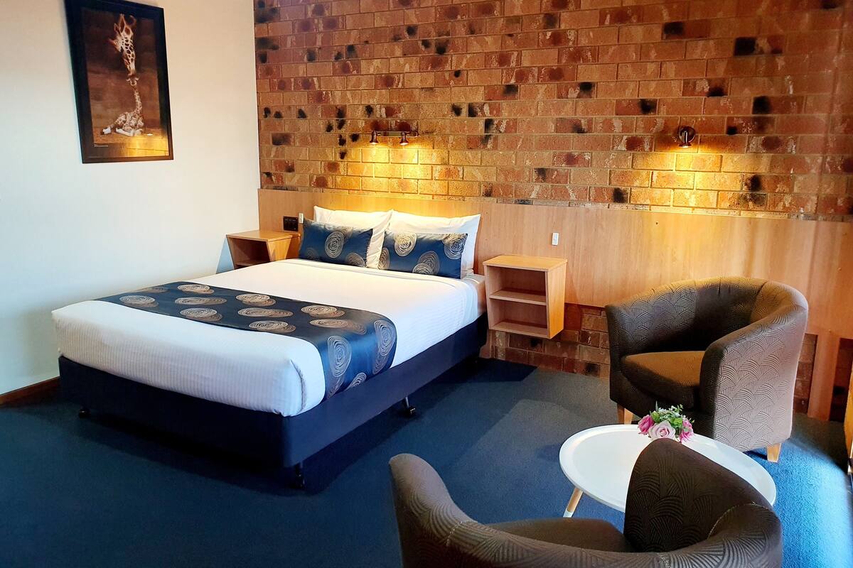 A hotel room with a large bed, a wooden nightstand, a chair, a small table with flowers, and a brick wall.