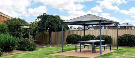 A covered patio area with a table and benches, surrounded by a well-maintained lawn and a wooden fence.