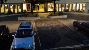 Un edificio moderno con múltiples ventanales y un acceso cubierto, rodeado de un estacionamiento con dos automóviles.