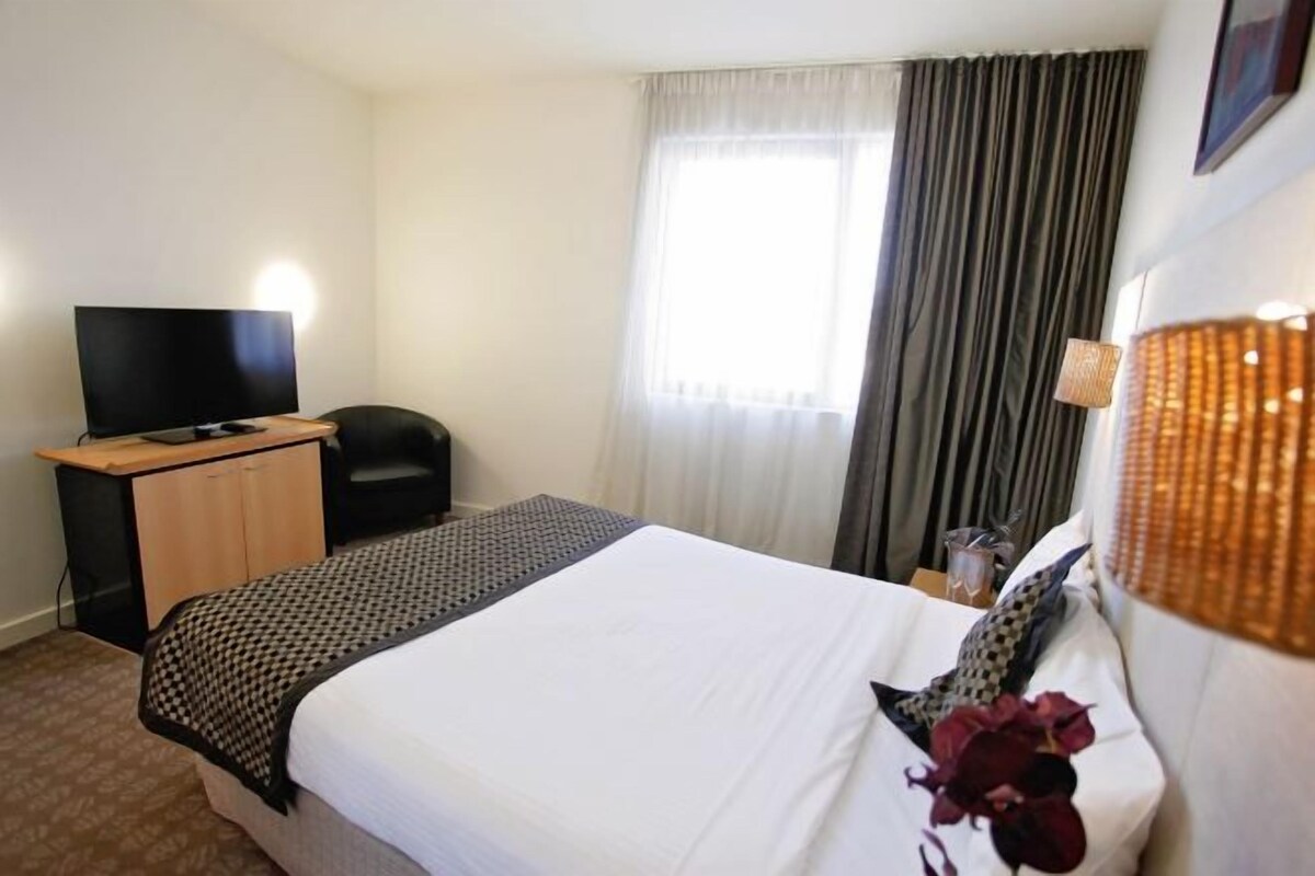 Habitación de hotel con cama, escritorio con televisor, una silla y ventana con cortinas.