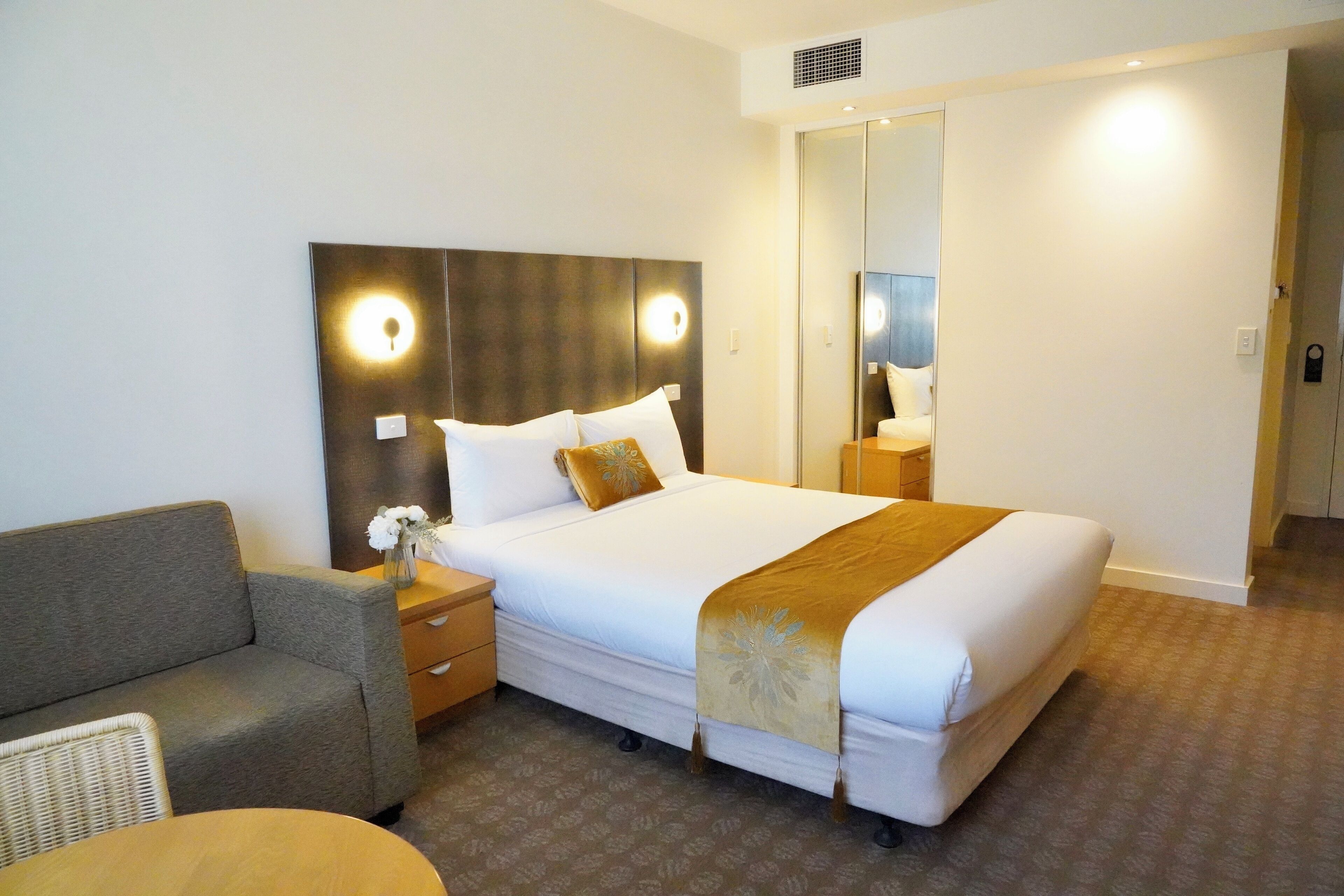 Ein Hotelzimmer mit einem großen Bett, einem Nachttisch mit Blumenvase, einem Sofa und einem Spiegel.