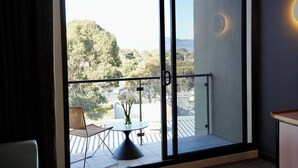 Un balcón con una mesa y sillas, vistas a árboles y una carretera, y un edificio con un elemento de iluminación circular.