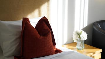 Cama bem arrumada com lençóis brancos e um travesseiro vermelho grande e texturizado. Um vaso transparente com flores brancas está sobre uma mesa de cabeceira de madeira.