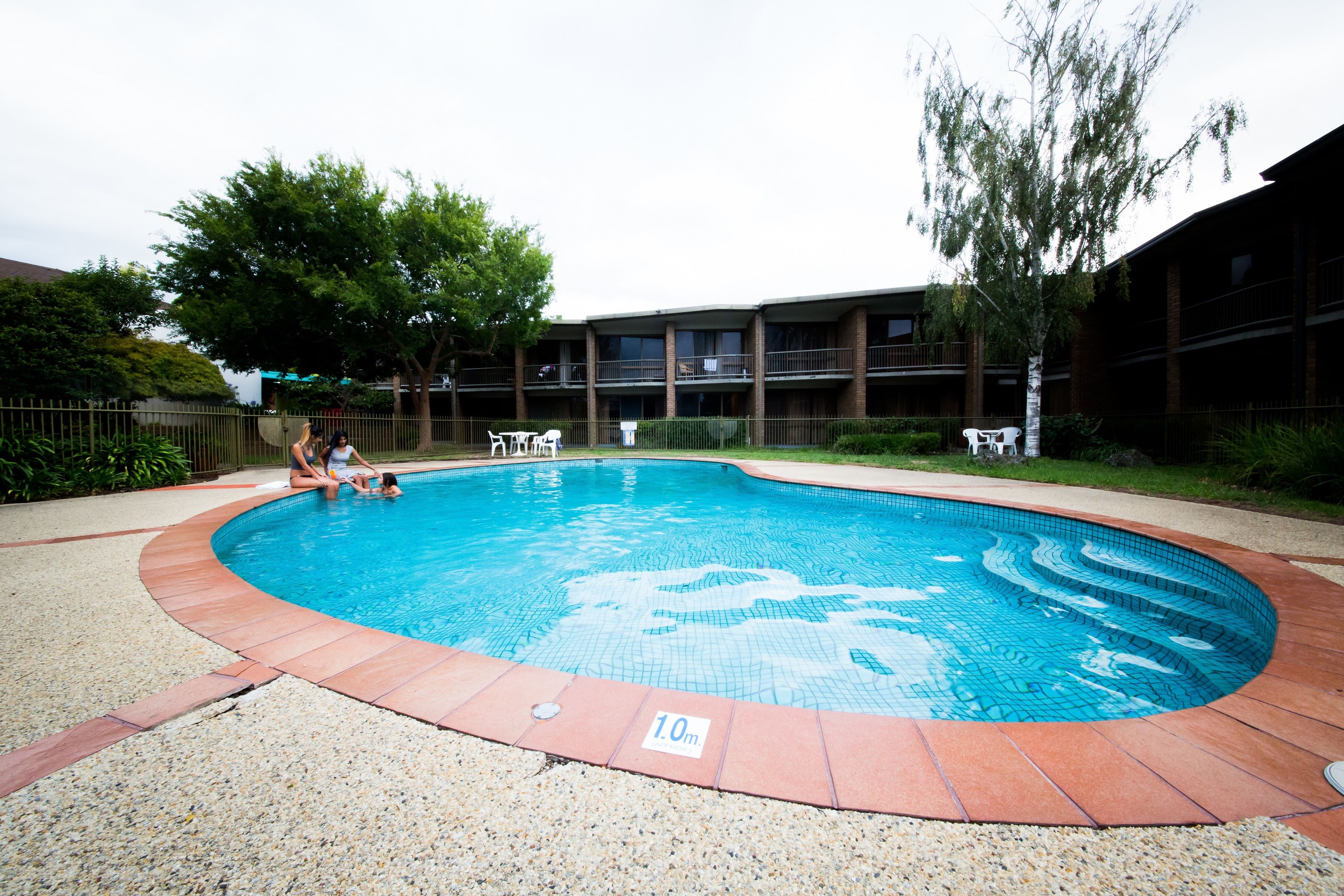 Una piscina con borde de azulejos, rodeada de grava y un edificio con varios balcones.