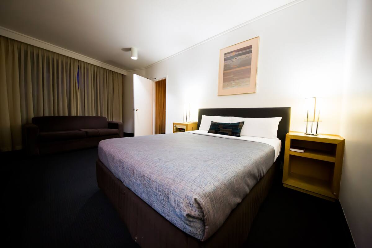 Siististi järjestetty hotellihuone, jossa on suuri sänky, yöpöytä lampun kanssa ja tummanvärinen sohva.